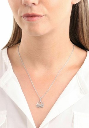 Collar de plata con un colgante de flor de loto, que presenta un intricado diseño calado sobre una fina cadena, llevado sobre una blusa blanca.