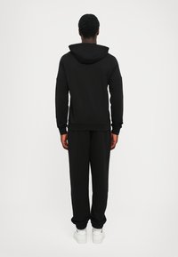 Felpa nera con cappuccio e pantaloni sportivi in tessuto morbido, dotati di cappuccio, polsini a costine e vestibilità comoda. Calzature includono sneakers bianche.