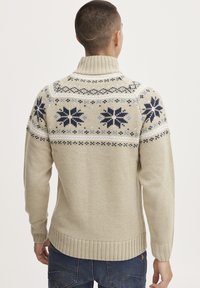 Beige neulottu neuletakki, jossa on korkea kaulus ja koristeellinen kuvio tummansinisellä ja vaaleansinisellä. Ribbauskainalot ja helma tuovat tekstuuria.