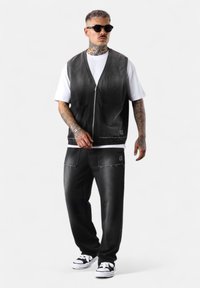 Schwarze Denim-Weste mit Reißverschluss und Taschen, über einem weißen T-Shirt getragen. Dazu passen abgenutzte schwarze Hosen und schwarz-weiße Sneaker.