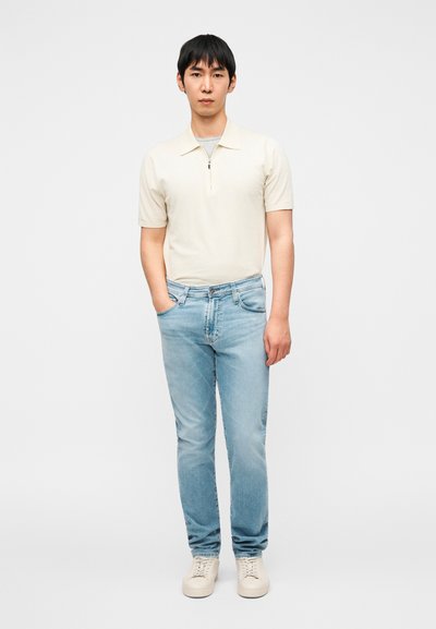 Ανοιχτό μπεζ κοντομάνικο πόλο με φερμουάρ, συνδυασμένο με ανοιχτό μπλε τζιν. Το υποκείμενο φορά λευκά παπούτσια και στέκεται μπροστά σε απλό φόντο.