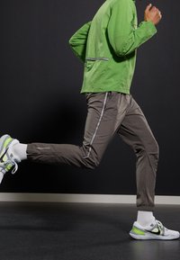 Veste légère verte avec accents réfléchissants, associée à un pantalon gris fuselé avec des bandes latérales, et des chaussures de sport blanches avec un détail vert.