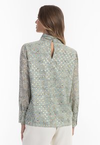 Blusa a fantasia floreale in verde menta, con maniche lunghe, colletto alto e dettaglio a oblò sulla schiena. Tessuto morbido e leggero.