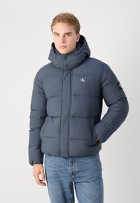 Mornarska puffer jakna z visokim ovratnikom, z zadrgo, horizontalnim šivom in logotipom na prsih. Nosita jo nad svetlo modrimi jeans hlačami.