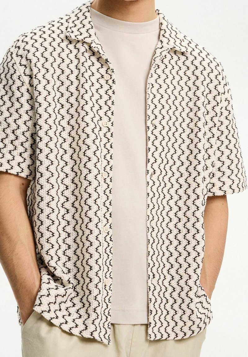 Kortärmad skjorta med krage och knappar i en krämfärgad nyans som har ett svart zigzag-mönster och en texturerad tyg. Bärs över en ljusgrå T-shirt.