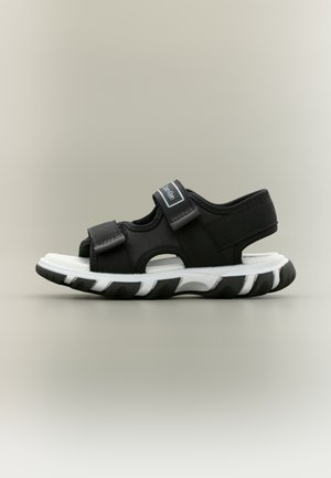 Sandale athlétique noire et blanche avec des sangles ajustables et une semelle épaisse, conçue pour le confort et un usage décontracté.