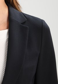 Tummansininen bleiseri, jossa on rakenteellinen kaulus, vartalonmyötäinen malli ja pehmeä kangas. Kerrostettu valkoisen teksturoituneen hupparin päälle. Minimalistiset muotoiluyksityiskohdat.