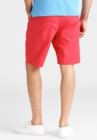 Red cotton shorts with a relaxed fit, two back pockets, and a smooth texture, paired with white sneakers.