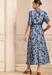 Rochie florală albastră cu mâneci scurtă făcute din puff, talie strânsă și lungime până la jumătatea gambei. Prezintă un model în culorile negru, roz și alb. Sandale cu toc maro.