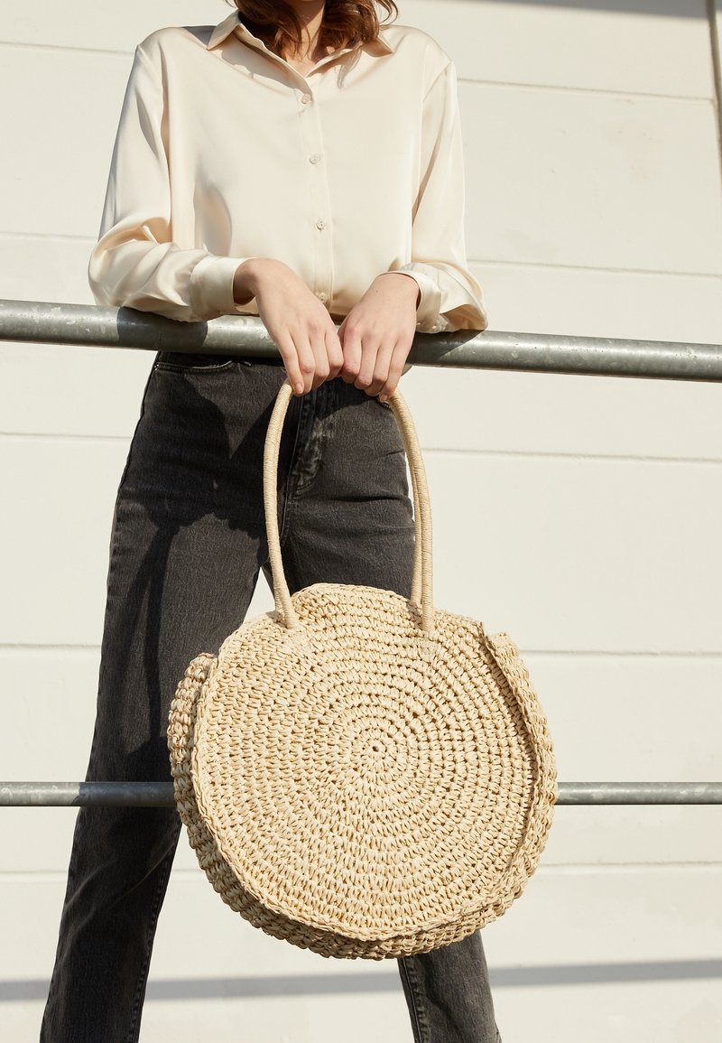 Rund halmhandväska med två korta handtag, med en vävd textur och en naturlig beige färg, hålls mot ett metallräcke.