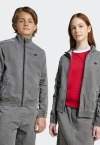 Des vestes en denim gris avec des poignets élastiques, comportant un petit logo. La fille porte une chemise rouge en dessous ; les deux ont un bas assorti.