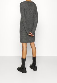 Šedé pletené šaty s dlouhými rukávy, kulatým výstřihem a žebrovanou strukturou. Nosené s černými robustními kotníkovými botami. Pozorováno ze zadní strany.