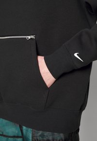 Svart sweatshirt med en ficka med dragkedja, ribbstickade ärmslut och en liten vit Nike-logotyp på ärmen. Tyget verkar vara texturerat och varmt.