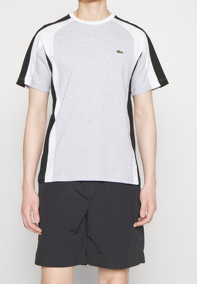 T-shirt de sport gris et noir avec des panneaux latéraux blancs, manches courtes, col rond et accent logo vert sur la poitrine. Short noir.