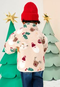 Pasztell rózsaszín, ünnepi mintákkal díszített bolyhos dzseki, amelyen Mikulás, rénszarvasok és fagyöngy minták láthatók. Élénk piros kötött sapkával párosítva.