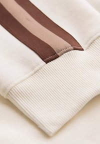Puño de sudadera blanca con una textura acanalada, acentuado con rayas horizontales en tela marrón y beige claro.