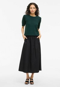 Top de punto verde de manga corta con un patrón texturizado, combinado con una falda maxi negra con cintura fruncida y bolsillos laterales. Sandalias negras de tiras.