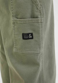 Pantalons cargo vert olive avec une poche latérale et une étiquette noire. Fabriqués à partir d'un tissu durable, avec des coutures piquées et une texture lisse.