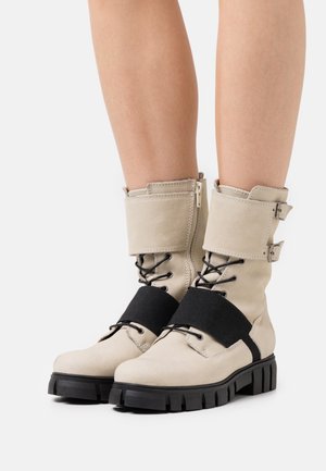 Botas de tobillo de ante beige con una tira elástica negra, parte delantera con cordones y cremalleras laterales. Suelas de goma negra gruesa con detalles texturizados.
