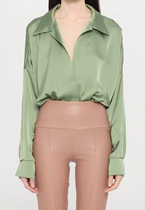 Blusa di raso verde con un ampio colletto e scollo a V profondo, abbinata a pantaloni nudi a vita alta e aderenti che mostrano una texture liscia.