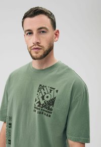 Hellgrünes Baumwoll-T-Shirt mit einem großen schwarzen Grafikdruck mit "PINK FLOYD" und Veranstaltungsdetails auf der Vorderseite. Kurze Ärmel und Rundhalsausschnitt.