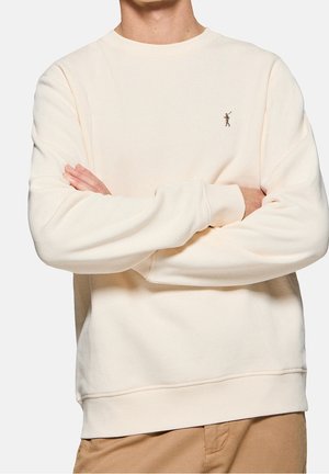 Person trägt einen cremefarbenen Pullover mit einem kleinen gestickten Polospieler-Logo auf der linken Brust und beigen Hosen, Arme verschränkt.