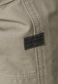 Béžová látka s mřížkovým vzorem, na které je černá nášivka G-Star Originals. Pro zvýraznění detailu jsou viditelné stehy podél okrajů.