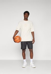 Kremfarget kortermet skjorte med grafikk på forsiden, sammen med svarte meshshorts og hvite treningssko, holder en oransje basketball.