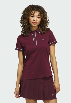 Young woman with curly hair wearing a maroon polo shirt and matching pleated skirt against a plain light background.