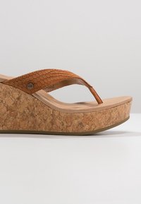 Sandál na klínu s corkovou platformou, světle hnědým stélkou a oranžovým texturovaným horním páskem, s logem na boku poblíž pásku.