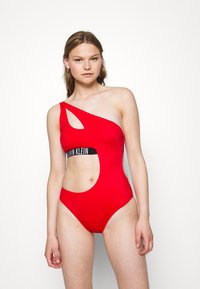 Červené jednodílné plavky s odvážným výřezem, jedním ramínkem a černým logem Calvin Klein obtočeným kolem pasu.