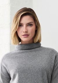 Junge Frau mit kurzen blonden Haaren, die einen grauen Rollkragenpullover trägt und mit neutralem Ausdruck direkt in die Kamera blickt.