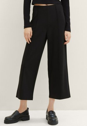 Frau trägt schwarze, weit geschnittene, knöchellange Hose, schwarzes langärmeliges Oberteil und schwarze klobige Plateau-Slipper, steht vor einem schlichten Hintergrund.