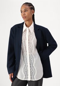 Marineblazer over en hvit skjorte med blonder hele veien nedover, kombinert med grå bukser. Glatte teksturer og strukturert design.