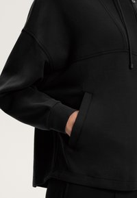 Fekete kapucnis pulóver laza fazonnal, kenguruzsebbel és állítható fűzőhúzóval. Sima textúra és minimális varratok.