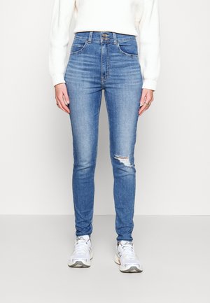 Kvinde iført højtsiddende, stramme blå jeans med et lille hul på højre knæ, hvid sweater og hvide sneakers, stående på en ensfarvet gulv.