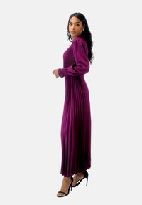 Robe longue plissée de couleur violet profond avec des manches longues et des poignets froncés. Portée avec des sandales à talons hauts noires. Texture de tissu lisse.
