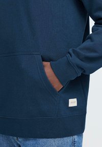 Felpa blu navy con vestibilità rilassata, polsini a coste, tasca frontale e una piccola etichetta in tessuto sul lato inferiore. Texture morbida.