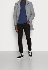 Manteau gris clair sur une chemise bleue ; pantalon noir ajusté ; baskets noires avec semelle blanche ; look superposé avec col distinct et détails de boutons.