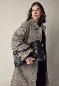 Černá kožená kabelka s dvojitými rukojetmi, držena proti hnědému kabátu s houndstooth vzorem, který má vykrojený límec a texturovanou látku.
