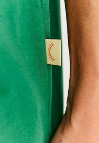 Zelená bavlnená tkanina s hladkým povrchom; obsahuje malú béžovú etiketu so zobrazením jednoduchého, stylizovaného dizajnu rias.