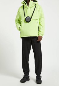 Hombre vestido con una chaqueta con capucha de color verde claro con bolsillos de cremallera negra, pantalones jogger negros y zapatillas negras, de pie con una mano en el bolsillo.