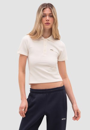 Kobieta z długimi blond włosami, ubrana w białą krótka koszulkę polo i czarne spodnie dresowe, stojąca na tle jednolitego jasnoszarego tła.