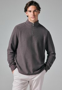 Pull-over à demi-fermeture éclair gris foncé en tissu texturé, avec un col haut et une coupe décontractée. Associé à un pantalon de couleur claire.