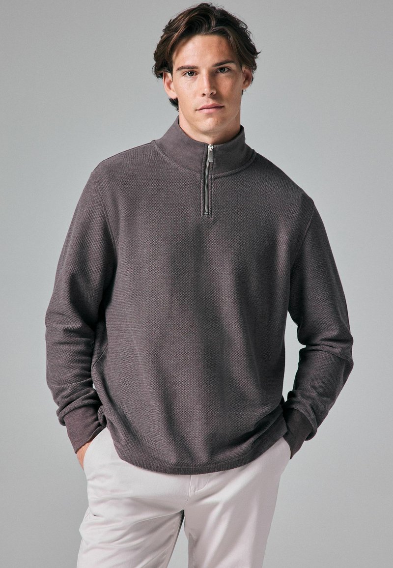 Dunkelgrauer Quarter-Zip-Pullover aus strukturiertem Stoff, mit hohem Kragen und lockerer Passform. Dazu helle Hosen.