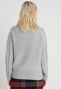 Světle šedý svetr vyrobený z pletené tkaniny, s pletenými detaily na rukávech a žebrovaným lemem. Nosený s kostkovanou sukní.