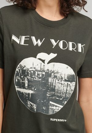 Ciemnozielona koszulka z nadrukiem "NEW YORK" nad białą grafiką miasta w kształcie jabłka oraz logo marki "SUPERDRY" blisko dołu.