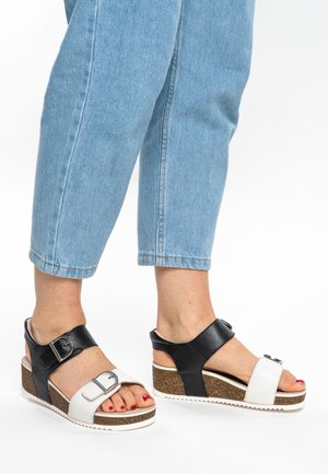 Black and white wedge sandals with buckle accents, cork platform, open toe, and textured straps. Worn with light blue cropped jeans.