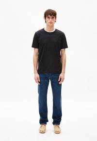 Schwarzes Baumwoll-T-Shirt mit Rundhalsausschnitt und einem dezenten, eingeprägten Logo, kombiniert mit blauen Jeans und beigefarbenen Mokassins.