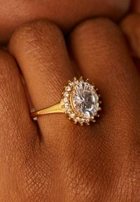 Gouden ring met een grote ovale heldere steen omringd door kleinere heldere stenen. De ring heeft een gladde band en een gepolijste afwerking.